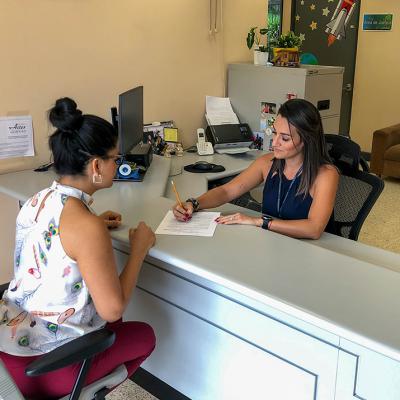 Se muestra a una mujer en una oficina llenando un documento y atendiendo a una señora